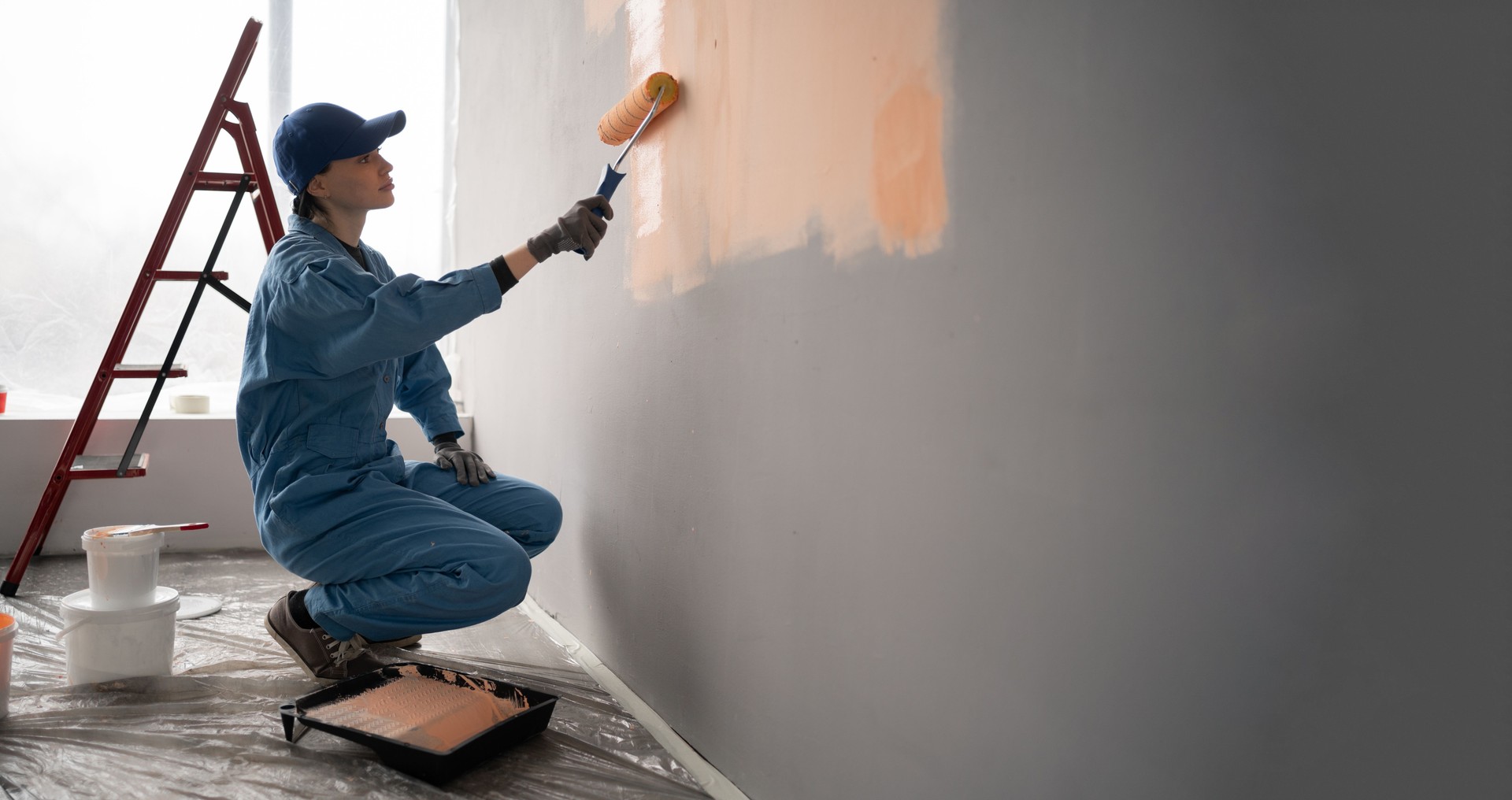female painter in overalls with cap and gloves painting a wall with paint roller, ladder and buckets on background. Copy space